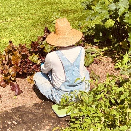 Kathleen S., 74, kneeling in her vegetable garden in a straw sun hat.